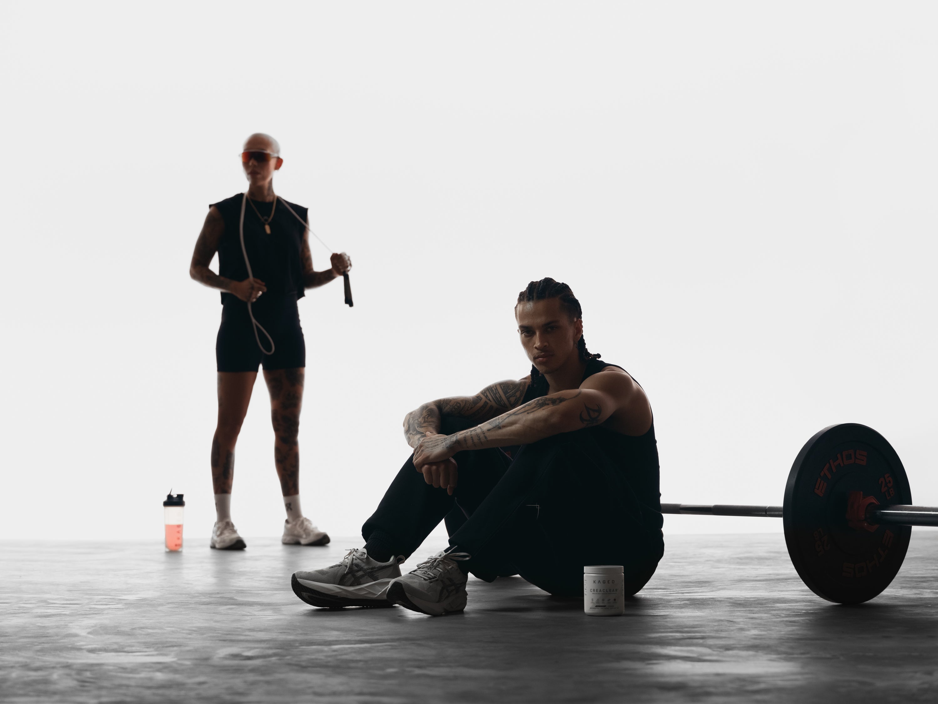 Two athletes in a bright studio, one seated by a loaded barbell and supplement tub, the other standing with a jump rope and shaker