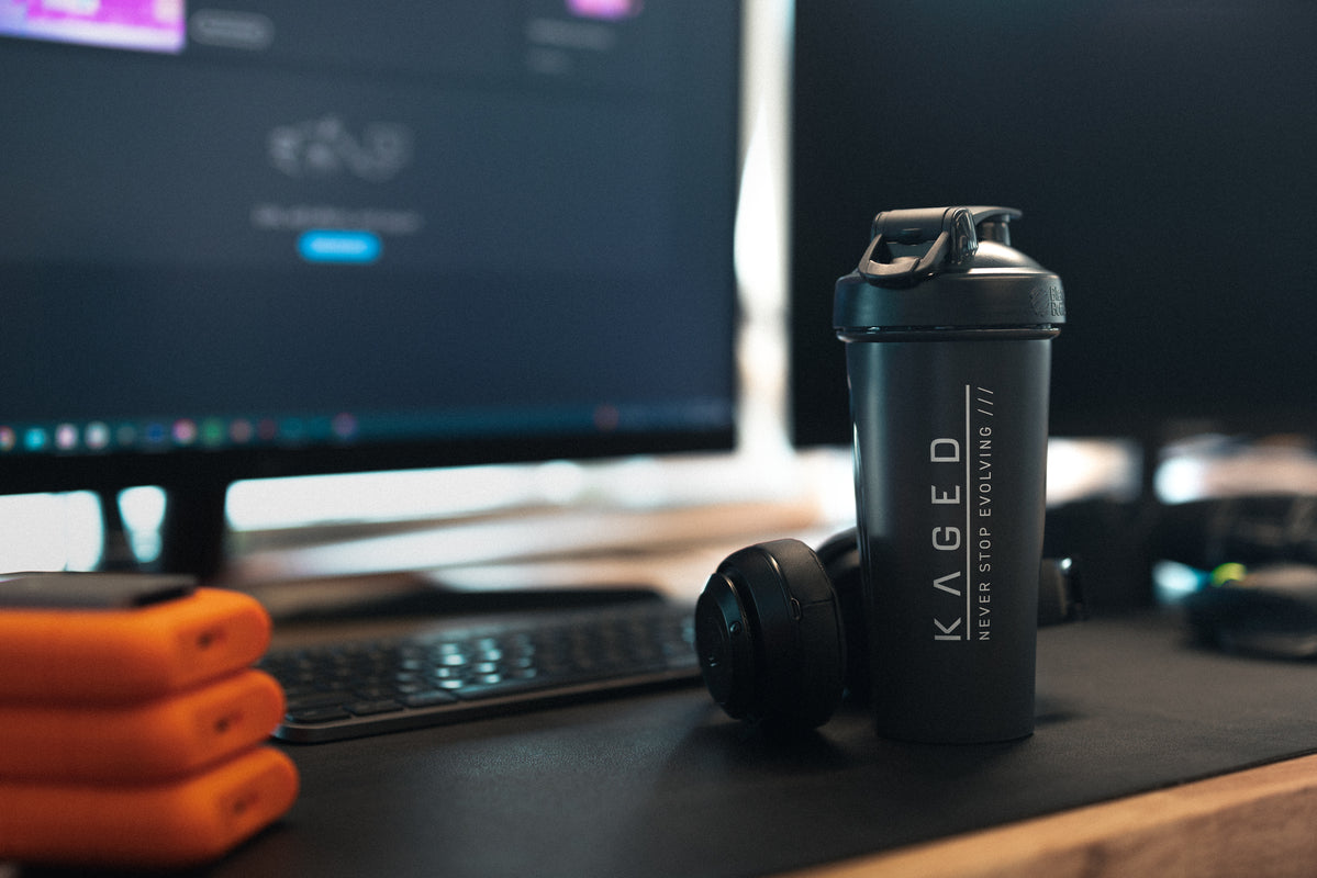 Three-quarter view of 28 oz Kaged Blender Bottle shaker with flip-top lid and KAGED logo on desk with headphones and monitor