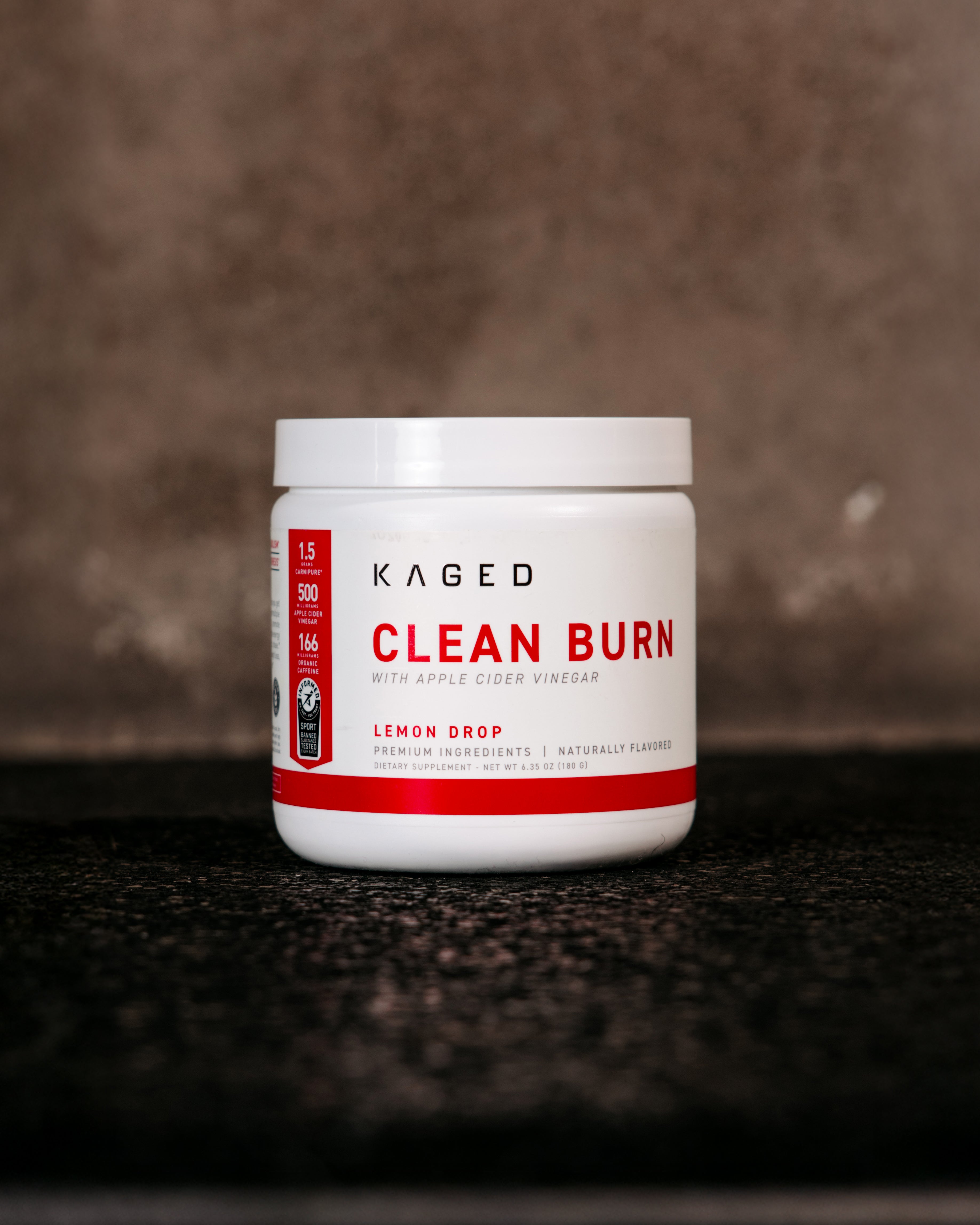 Front view of Kaged Clean Burn powder jar showing "Clean Burn with apple cider vinegar" label and Lemon Drop flavor on a dark textured surface