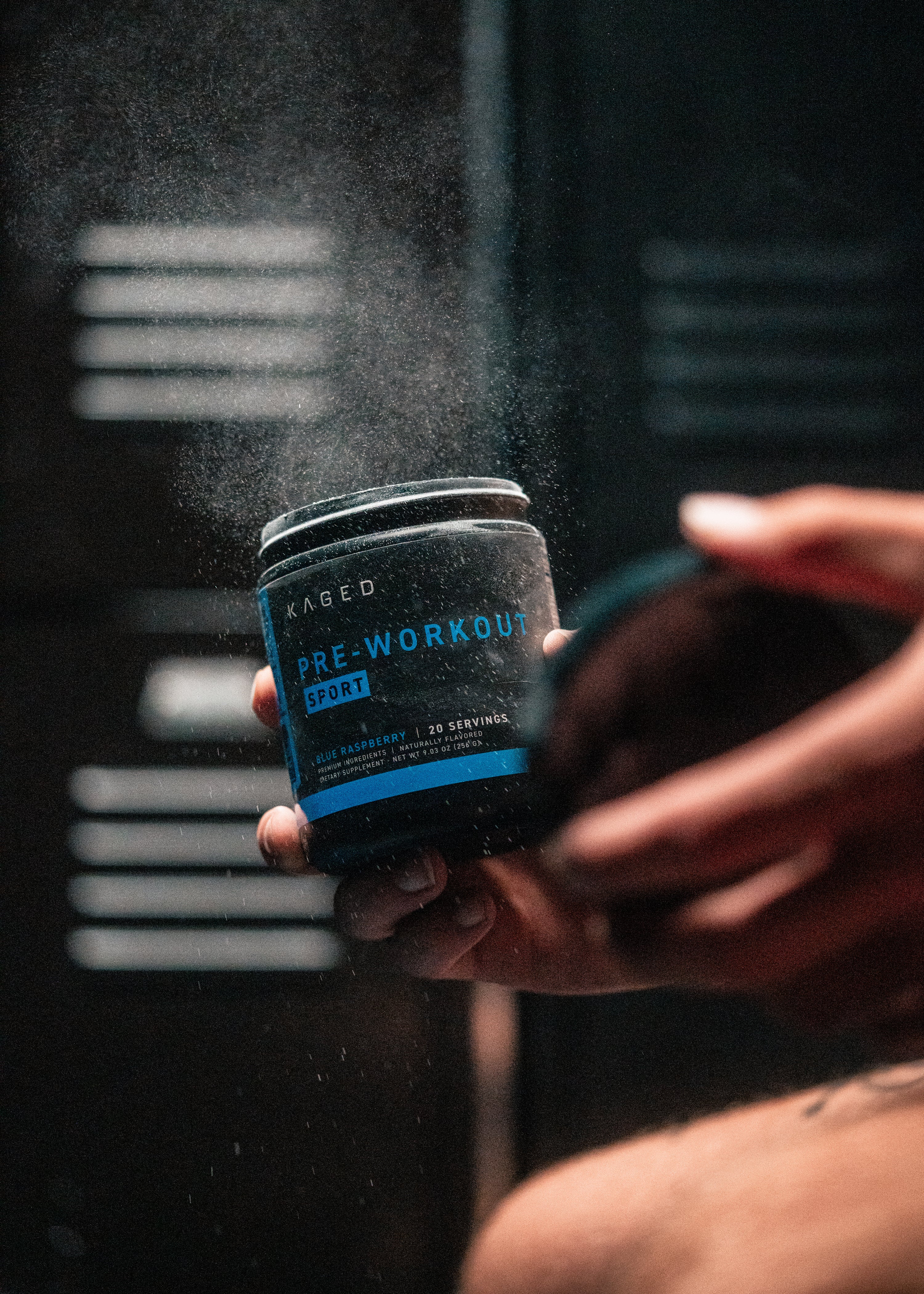 Close-up of KAGED Pre-Workout Sport tub held in hand showcasing blue label and powder mist in a dark gym setting