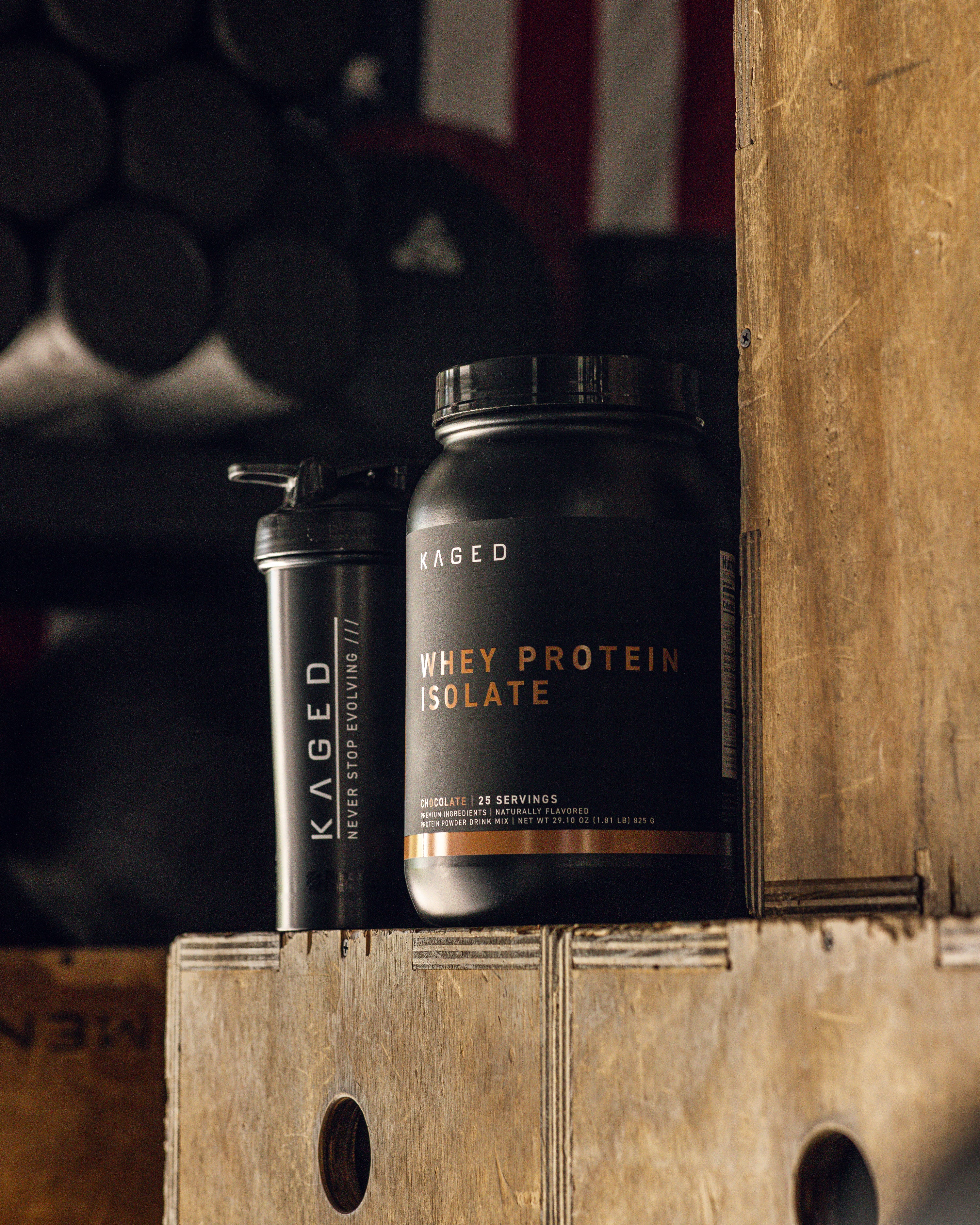 Front view of Kaged Whey Protein Isolate tub and shaker on wooden gym boxes showing the Whey Protein Isolate label