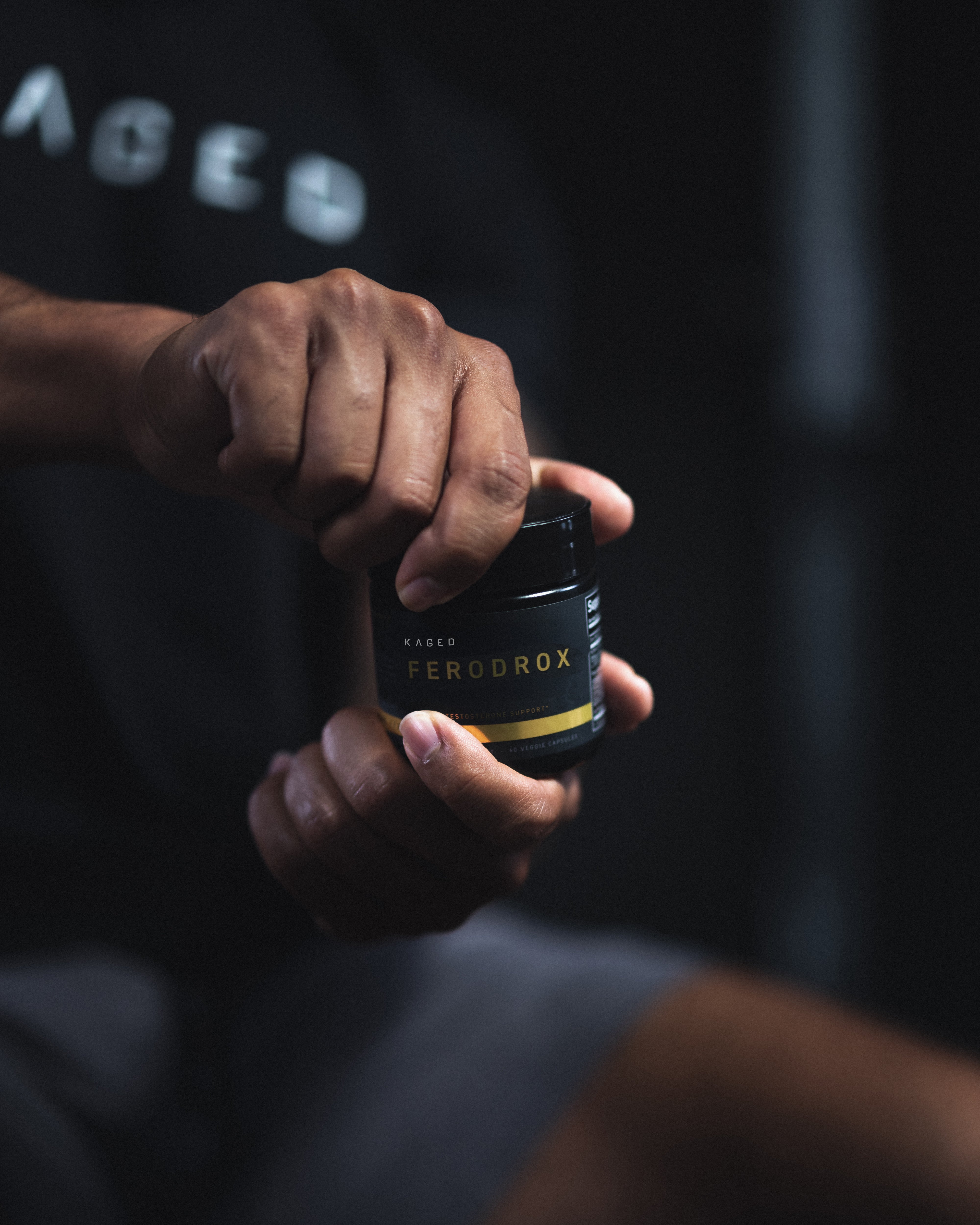 Close-up of Ferodrox supplement jar held and being opened by hands, showing the KAGED label against a dark blurred background