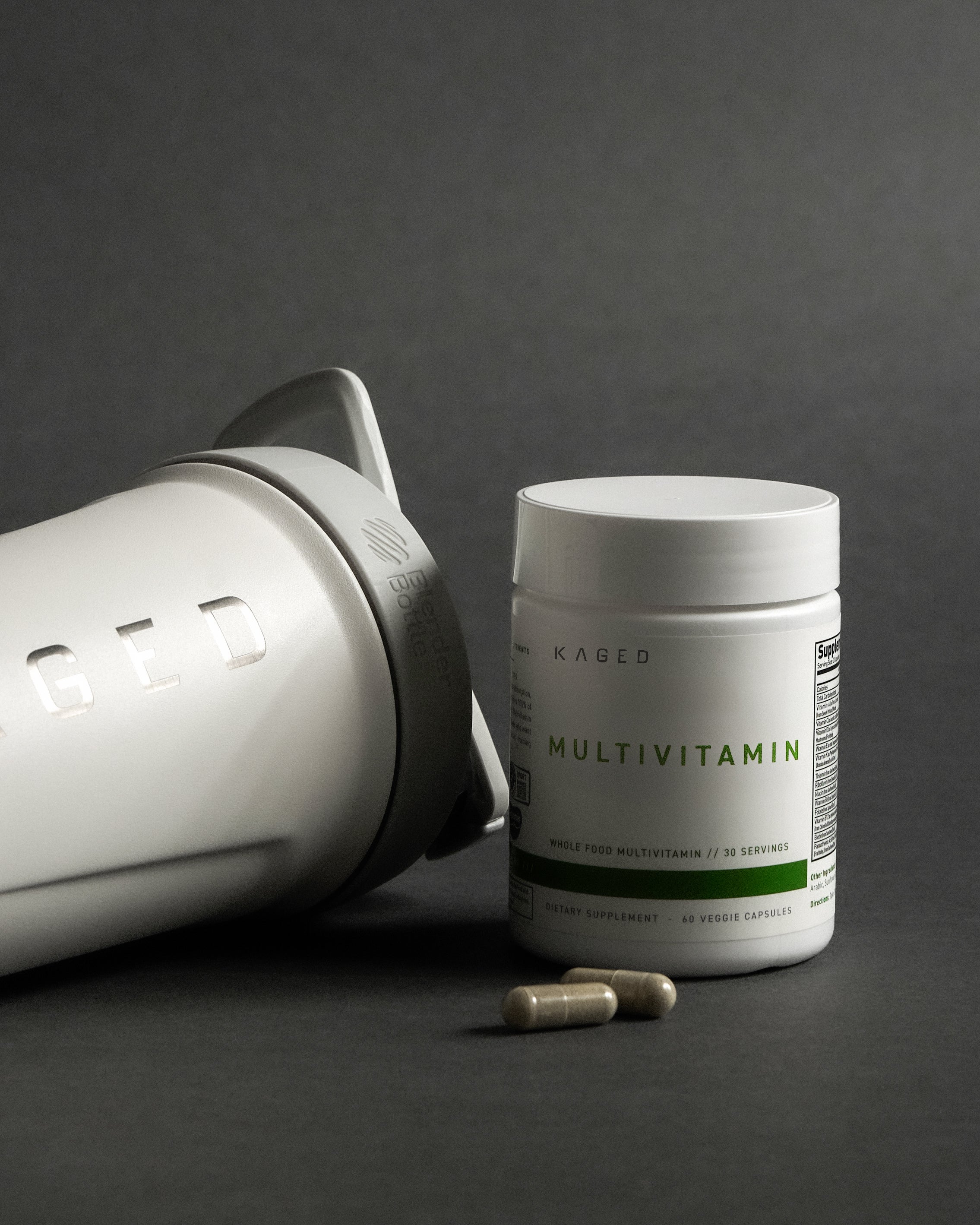 Close-up of Kaged Multivitamin bottle showing MULTIVITAMIN label and two capsules beside a Kaged shaker on gray background