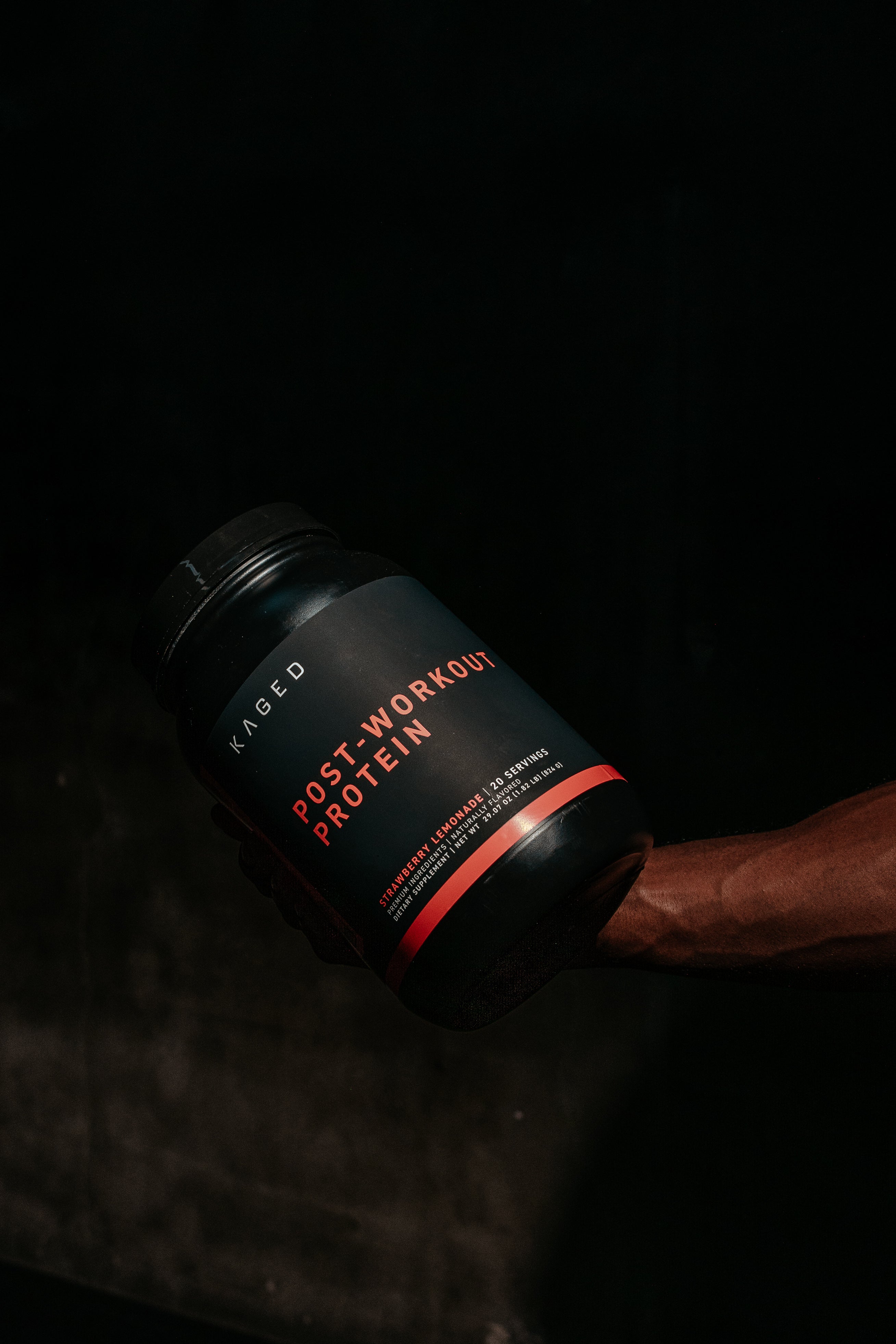 Angled close-up of Re-Kaged Post-Workout Protein tub showing front label reading POST-WORKOUT PROTEIN against a dark background
