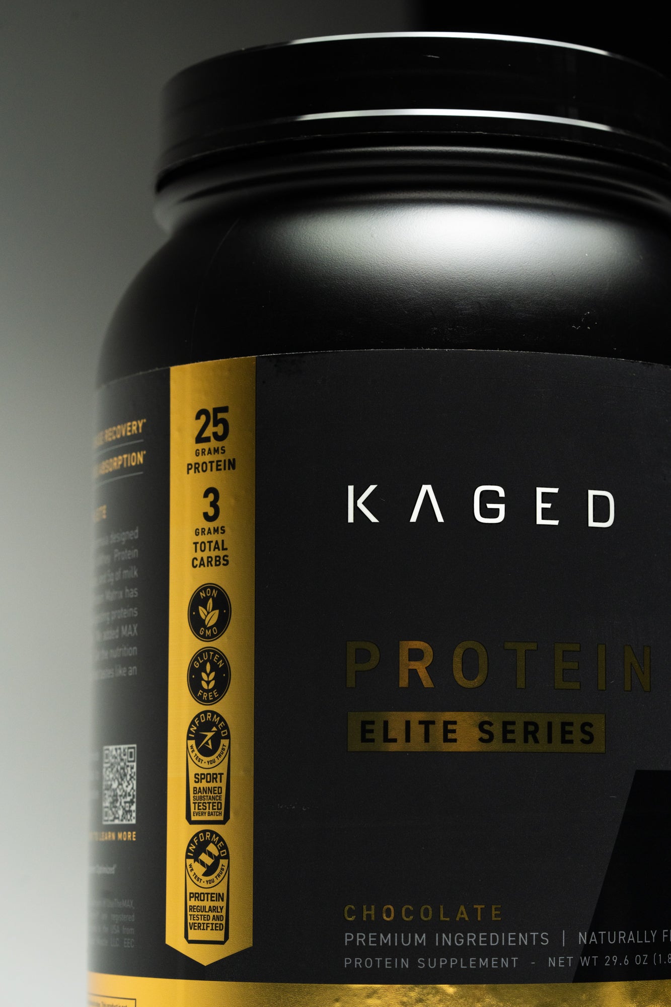 Close-up of Kaged Protein Isolate Elite chocolate tub showing KAGED logo and gold nutrition strip stating 25 grams protein on a dark background