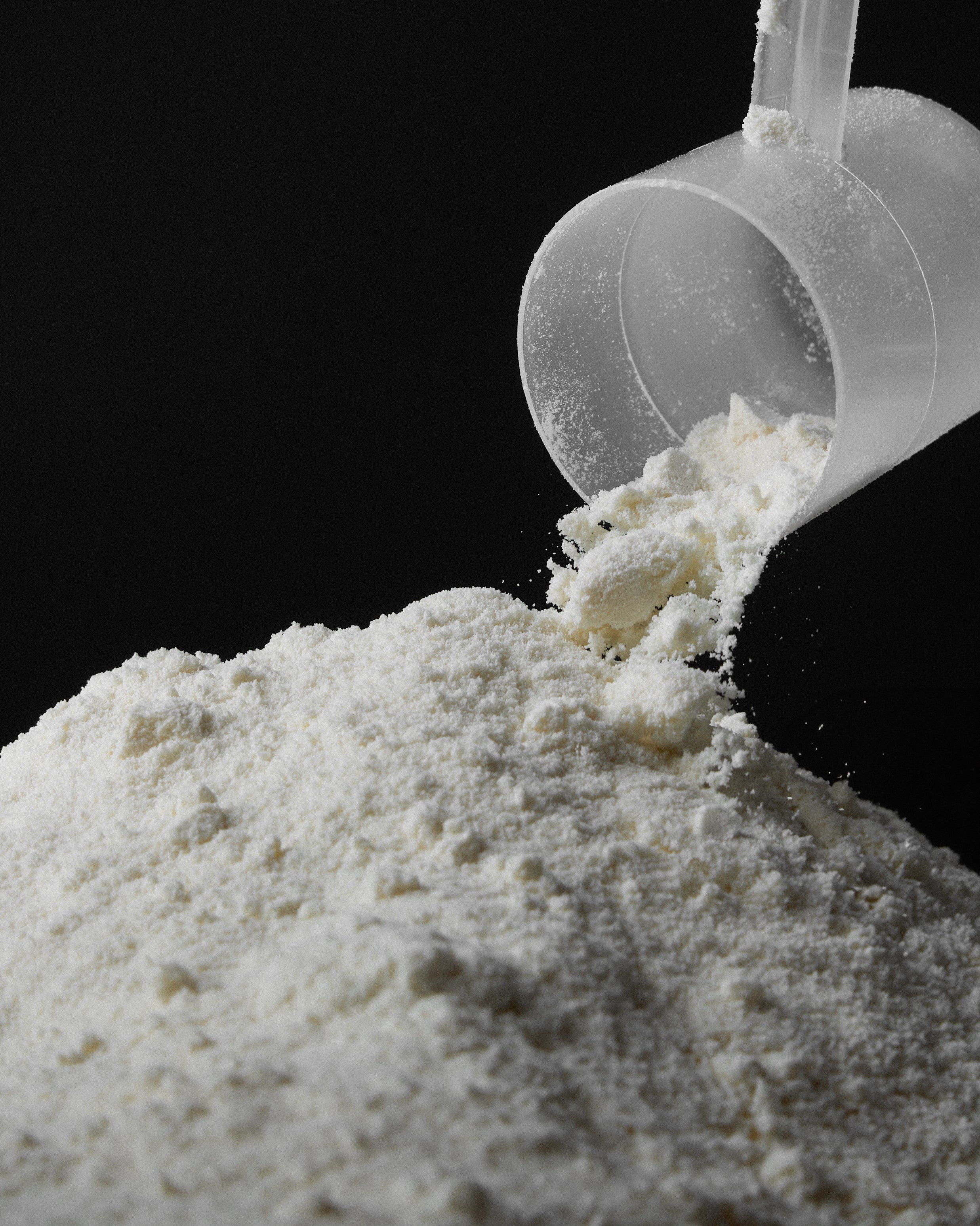 Close-up of a clear scoop pouring white protein powder onto a pile against a black background