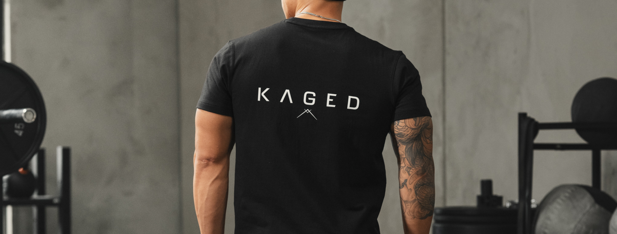 Back view of black Ascent Tee, slim fit, showing white KAGED logo across the back in a gym setting
