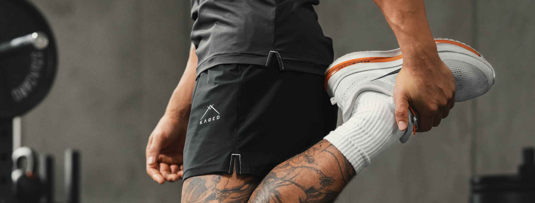 Close-up side view of Ascent Performance shorts showing KAGED logo and split hem while model stretches in gym