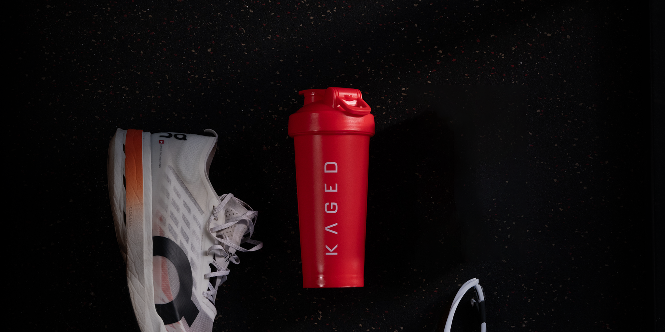 Top-down view of crimson 28oz shaker bottle showing vertical KAGED logo and flip-top lid on speckled gym floor beside shoe