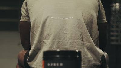 Rear view of person on bench wearing beige shirt reading FUEL YOUR EVOLUTION, blurred Kaged pre-workout tub in gym
