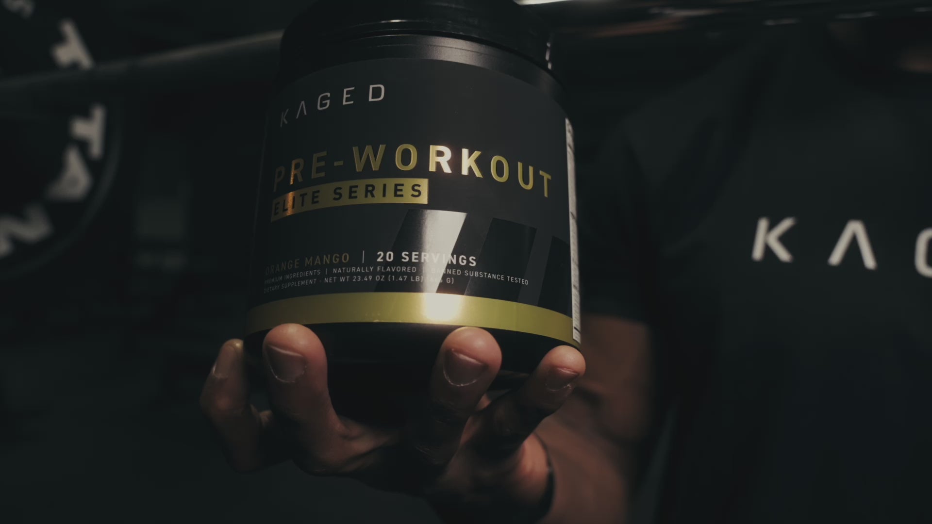 Hand holding a KAGED pre workout tub in a low-light gym setting, moody close-up showing Pre-Workout label