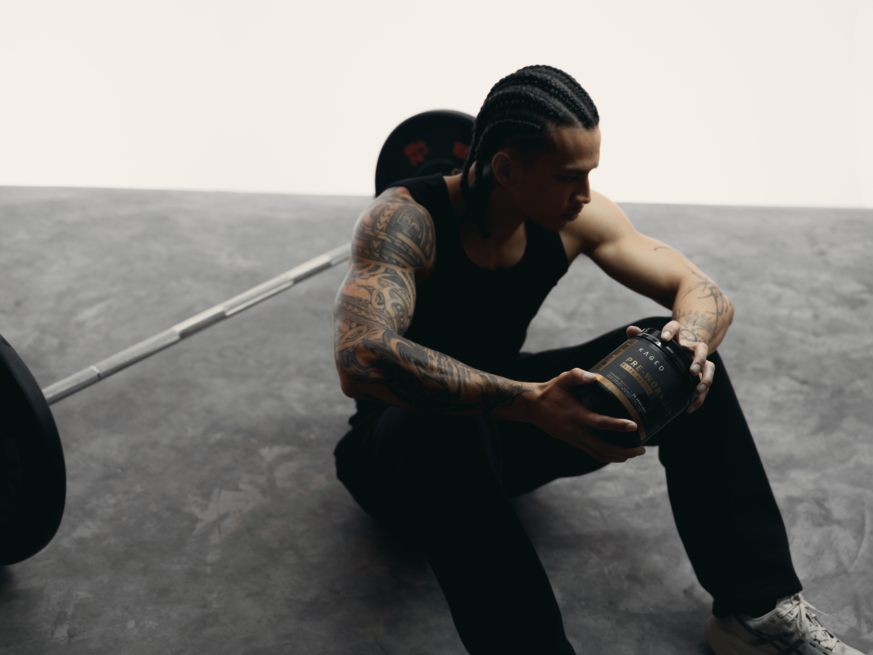 Athlete sitting on a gym floor holding a KAGED Pre-Workout tub beside a loaded barbell